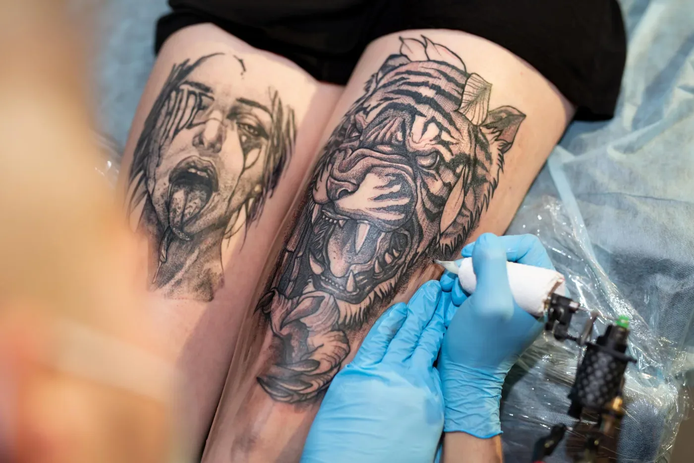 Gloved artist working a tattoo machine over a client's leg mid-session
